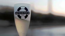Champagne flute with Vedettes du Pont Neuf logo, against a blurred background of the Seine and Eiffel Tower at sunset