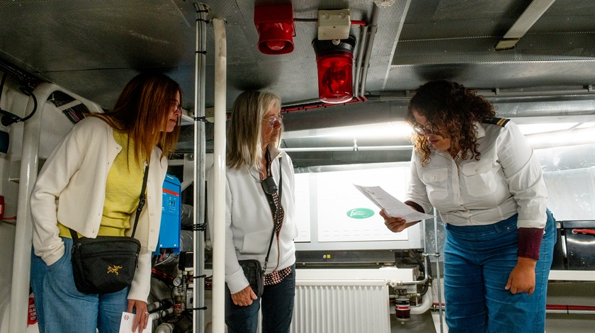 Une membre de l’équipage consulte un document devant deux visiteuses dans la salle des machines d’un bateau.