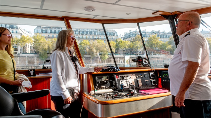 Deux visiteuses échangent avec un membre de l’équipage devant le tableau de bord d’un bateau, dans la cabine de pilotage.